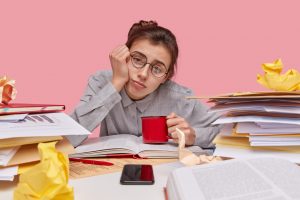 Chica joven estudiando con dificultades de concentración, rodeada de libros en representación de la sintomatología TDAH. Tiene expresión de cansancio, sostiene una taza de café entre en un entorno desordenado.