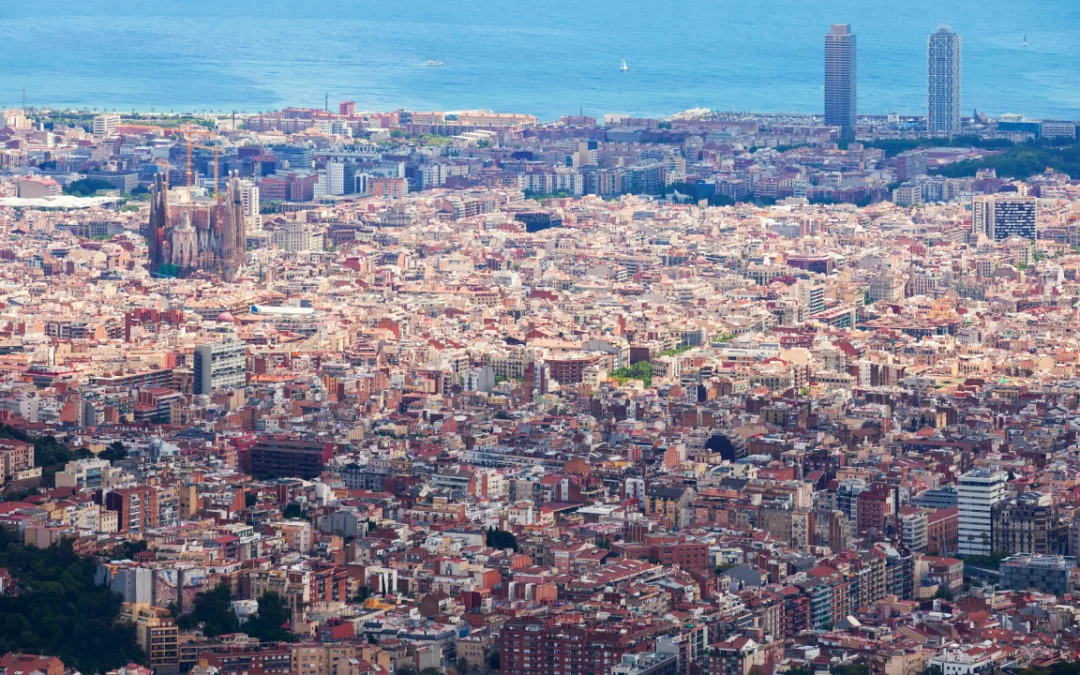 High-angle aerial view of Barcelona showing dense urban building blocks, the Sagrada Família, and the coastline, illustrating urban density and spatial pressure.