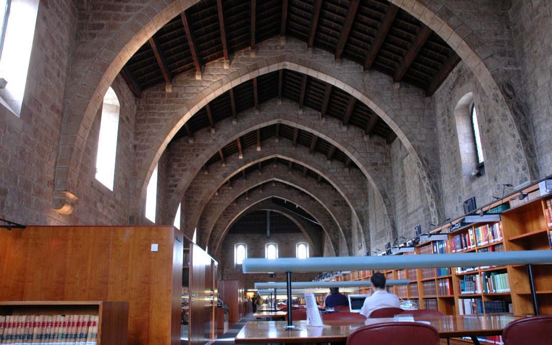 Nau de tramuntana de la Biblioteca de Catalunya amb arcs de pedra gòtics, taules d'estudi i prestatgeries de fons documental.