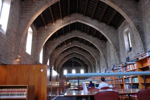 Nau de tramuntana de la Biblioteca de Catalunya amb arcs de pedra gòtics, taules d'estudi i prestatgeries de fons documental.